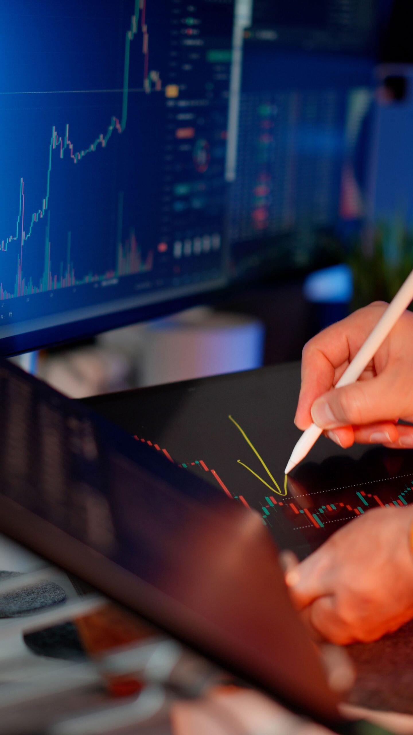 Person using a stylus on a tablet for stock market analysis, with charts on a monitor.